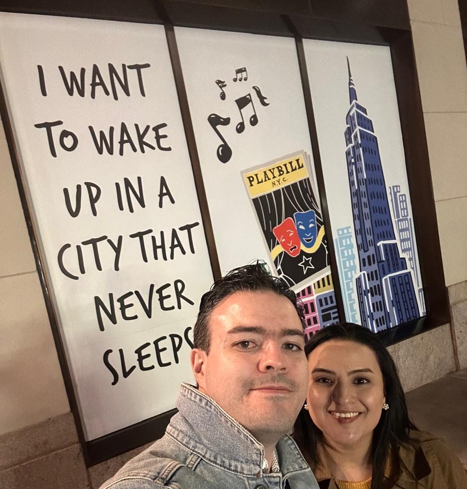 Two people outside a painted Playbill mural in New York, under a line reading "I want to wake up in a city that never sleeps".