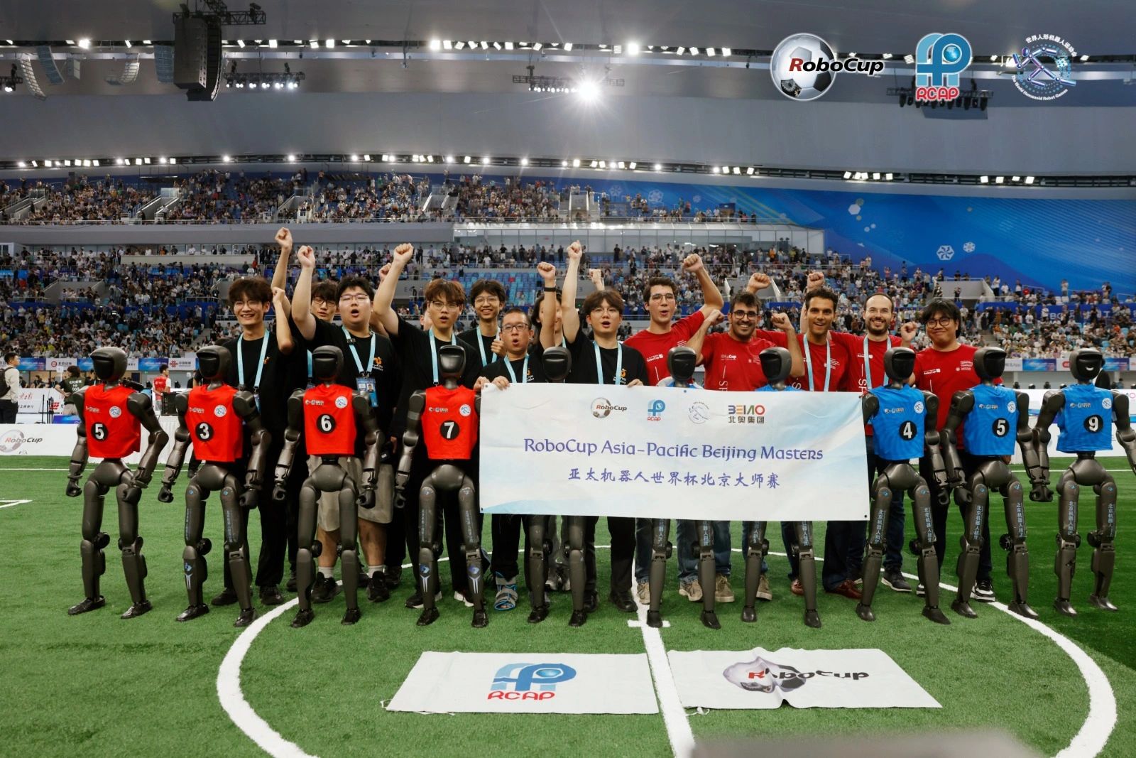 Equipos de Europa y Asia celebrando con sus robots humanoides bajo la manta del RoboCup Asia-Pacific Beijing Masters, en un National Speed Skating Oval lleno.