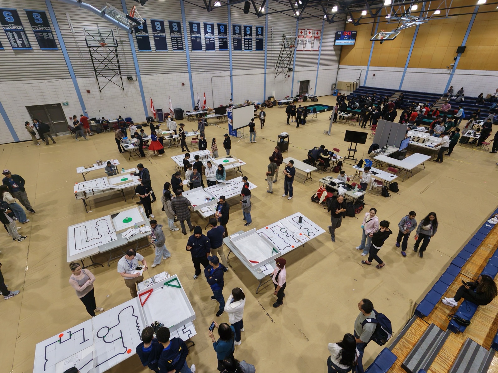 Vista aérea de la sala de competencia en Sheridan College, en Brampton: filas de canchas de Rescue Line de RoboCupJunior sobre mesas, con estudiantes y mentores trabajando alrededor.