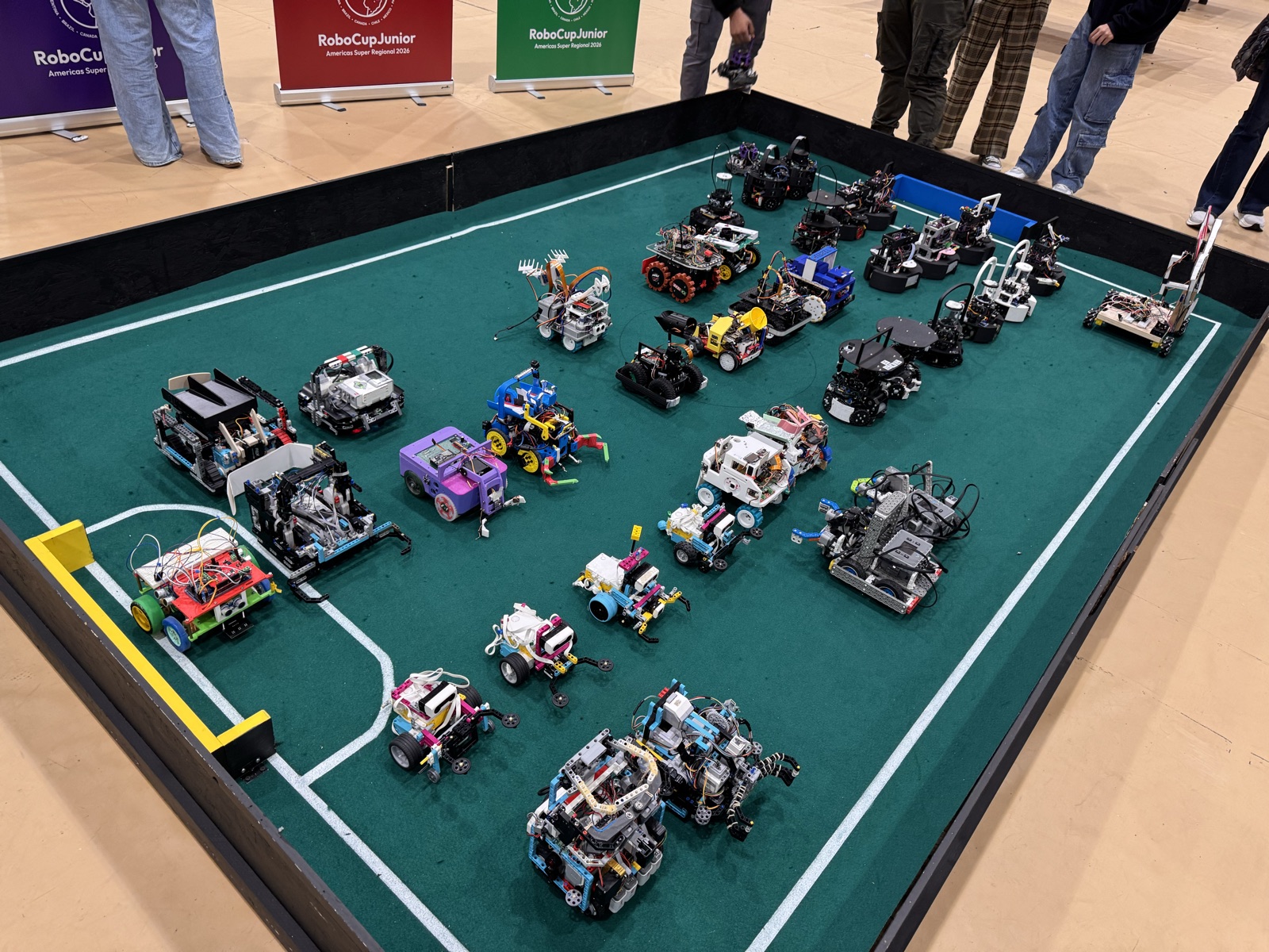 Decenas de robots de fútbol y de rescate construidos por estudiantes, alineados sobre una cancha verde de RoboCupJunior, con los banners de las ligas detrás.