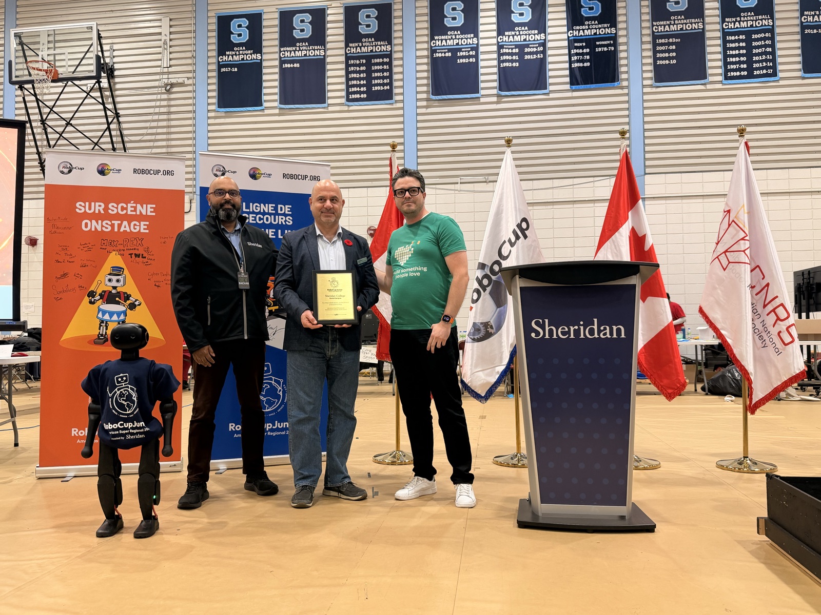Tres personas de pie junto al podio de Sheridan en el gimnasio, con los banners de la competencia y las banderas detrás; la persona del centro sostiene una placa.